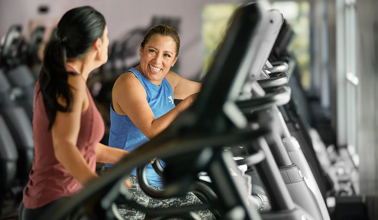 Two women smiling and chatting while working out on elliptical machines at the gym